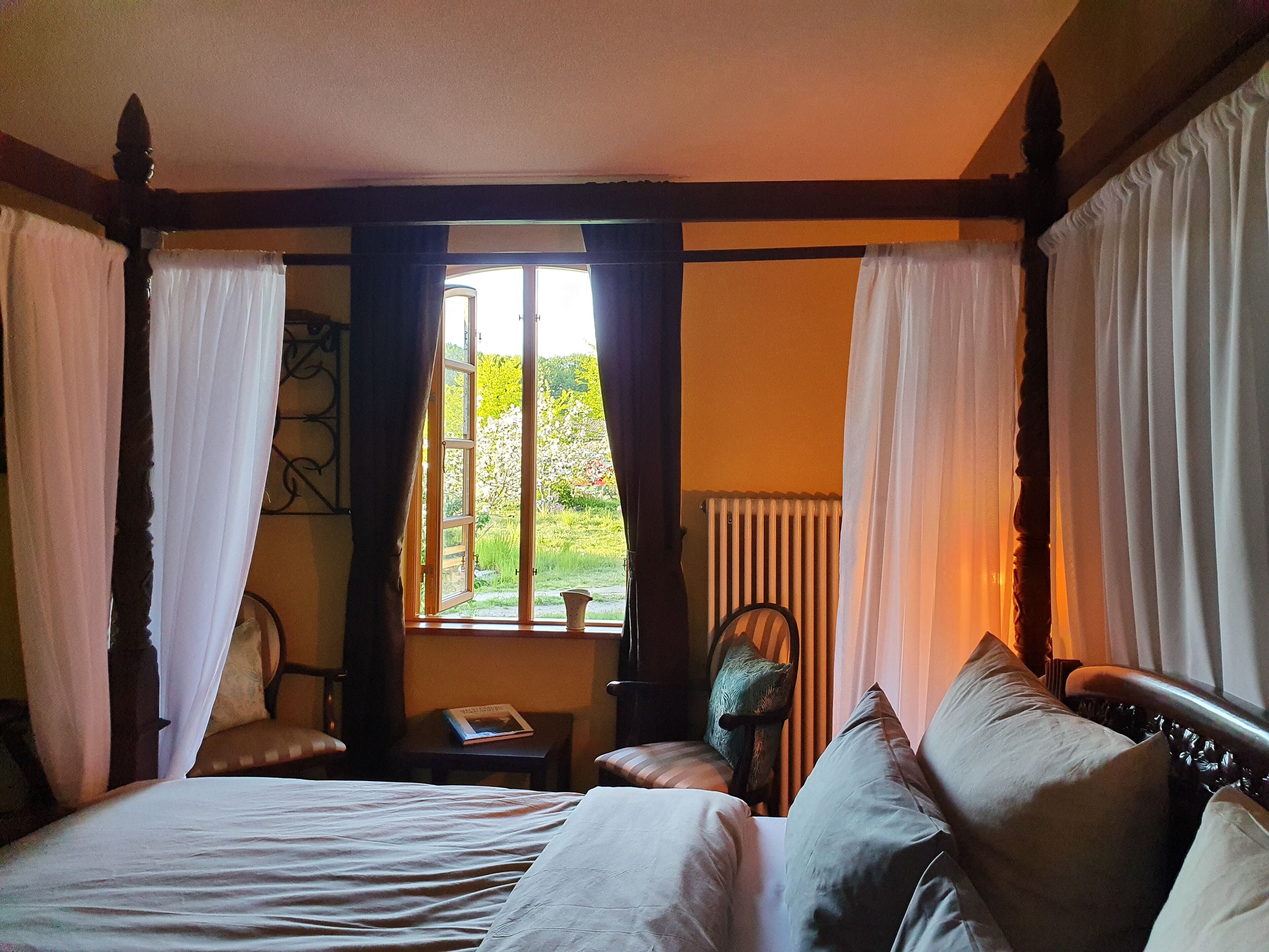 Ein Schlafzimmer mit einem großen Bett, einem Sessel, einem kleinen Tisch und einem Fenster mit Vorhängen.