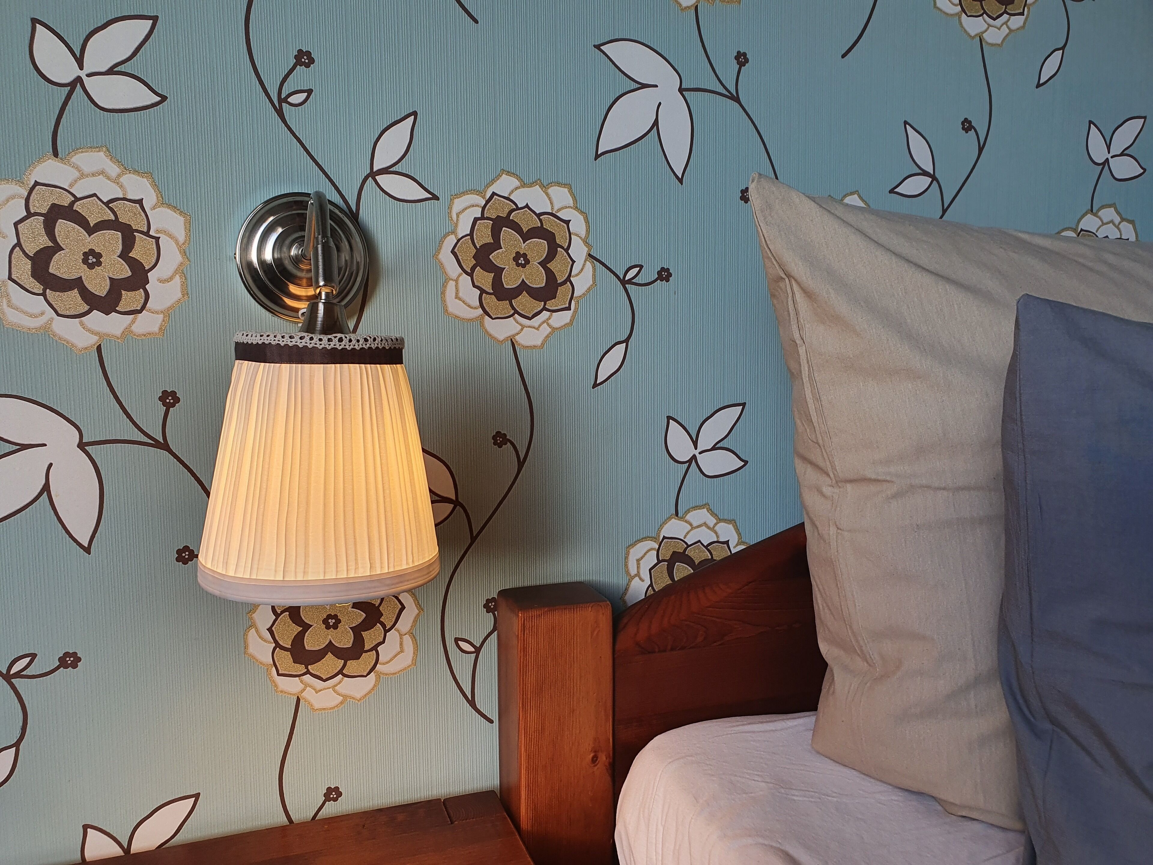 A bedroom with floral wallpaper, a bedside lamp, a wooden nightstand, and a bed with pillows.