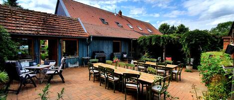 Eine Terrasse im Freien mit Backsteinböden, Holztischen und -stühlen, einem Grill und einer überdachten Sitzgelegenheit mit blauem Markisenstoff.