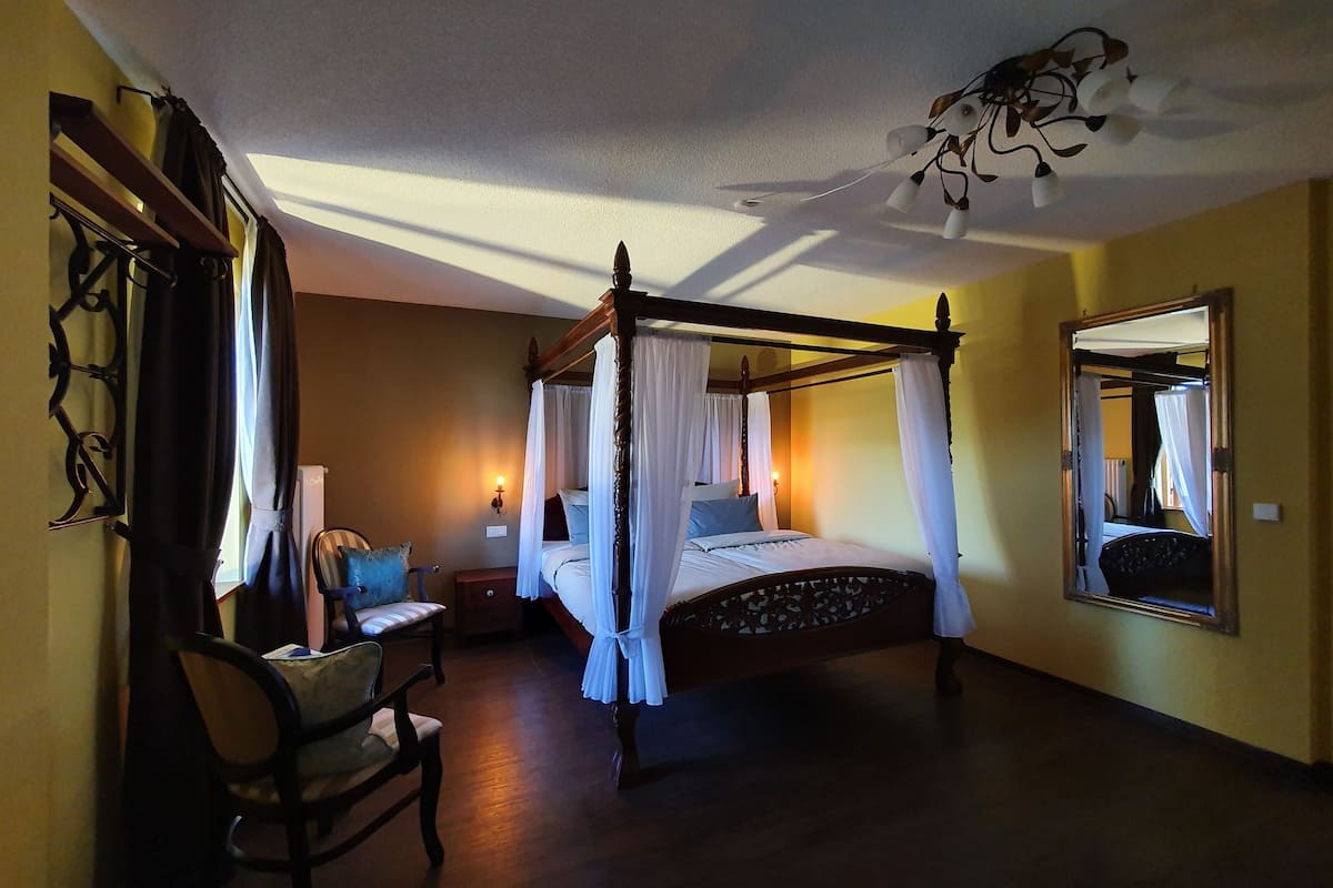 Un lit à baldaquin à quatre poteaux avec une literie blanche, une table de chevet en bois avec une lampe, une chaise avec un coussin et un miroir accroché au mur.