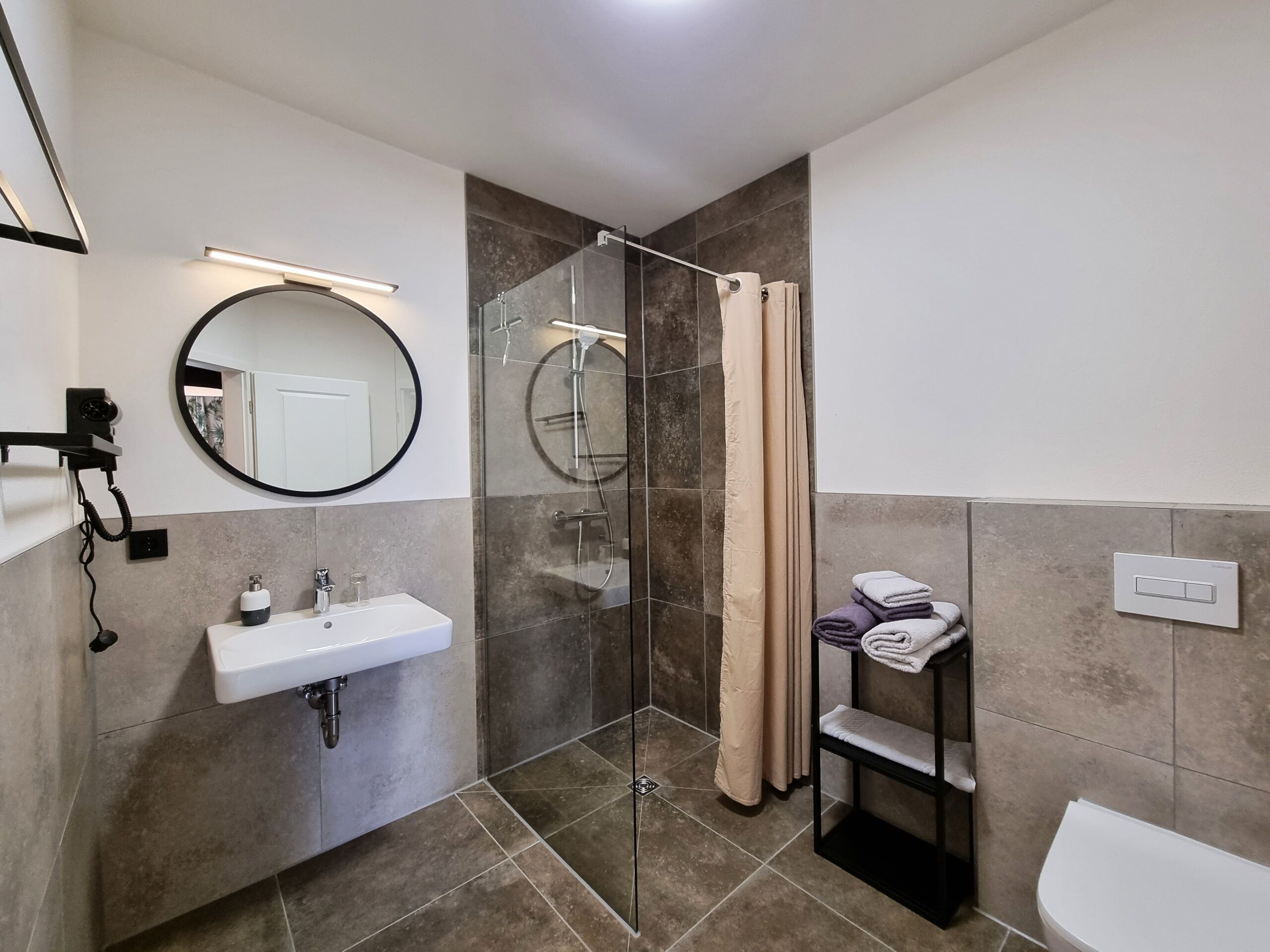 A modern bathroom with a glass-enclosed shower, a round mirror, a white sink, and a towel rack.