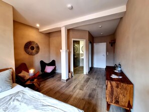 Un dormitorio con cama, una mesita de noche, una silla, una mesita auxiliar de madera, un espejo montado en la pared y una puerta que da al baño.