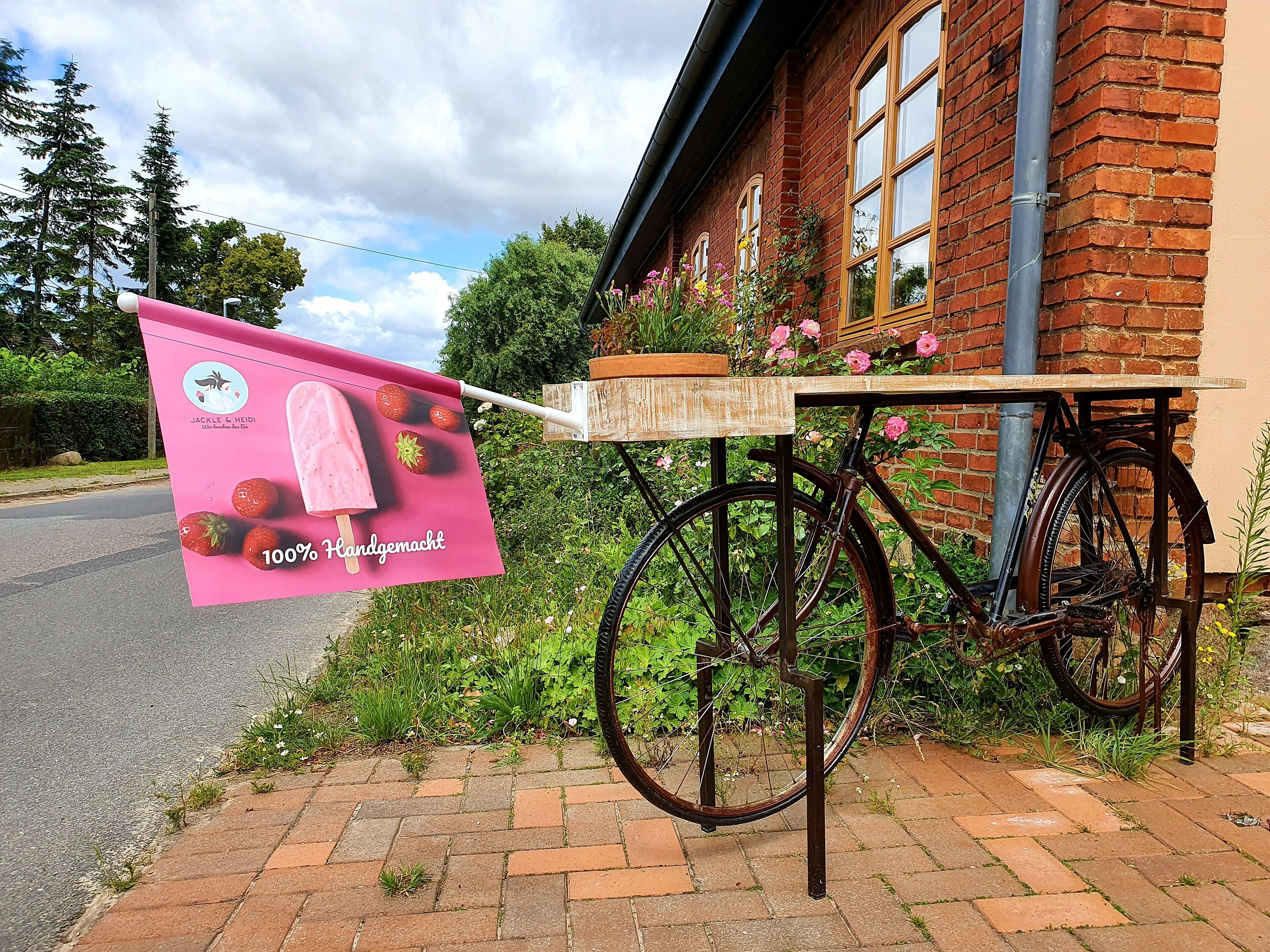 Un vélo muni d’une table fixée, garé devant un bâtiment en briques doté de fenêtres et d’une jardinière.