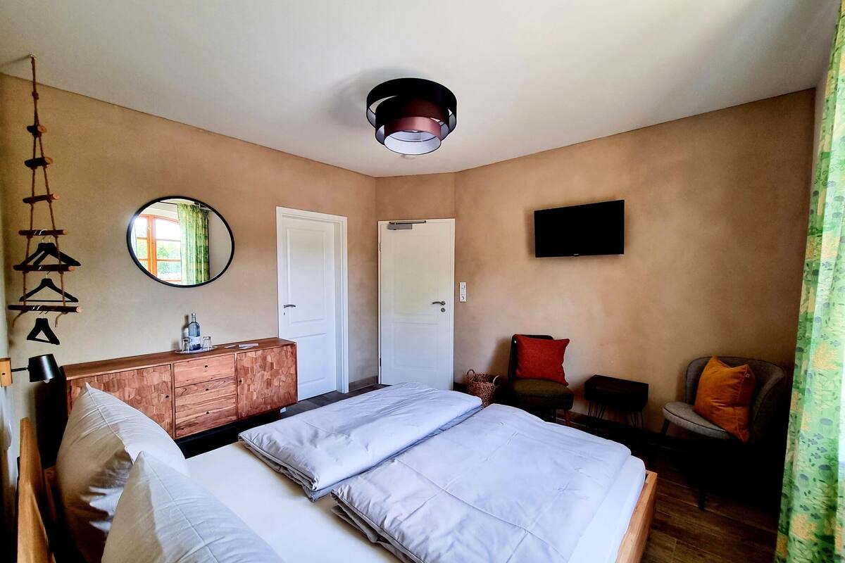 Une chambre à coucher avec un lit, une commode en bois, un miroir, une télévision et une chaise avec des coussins.