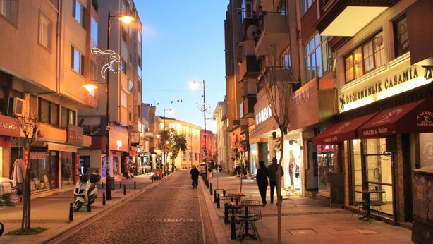 A cobblestone street lined with buildings, some with signage indicating shops and cafes. - Degirmenlik Casamia Suite (Çanakkale)