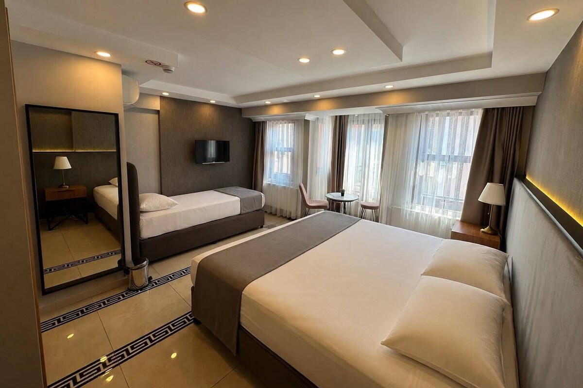 A hotel room with a large bed, a bedside table, a TV, a small table with chairs, and a window with curtains.
