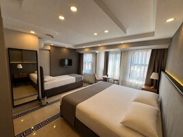 Habitación de hotel con una cama grande, una mesita de noche, un televisor, una pequeña mesa con sillas y un ventanal con cortinas.