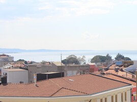 Un paisaje urbano costero con tejados de tejas color terracota, edificios con balcones y vista al mar y montañas distantes.