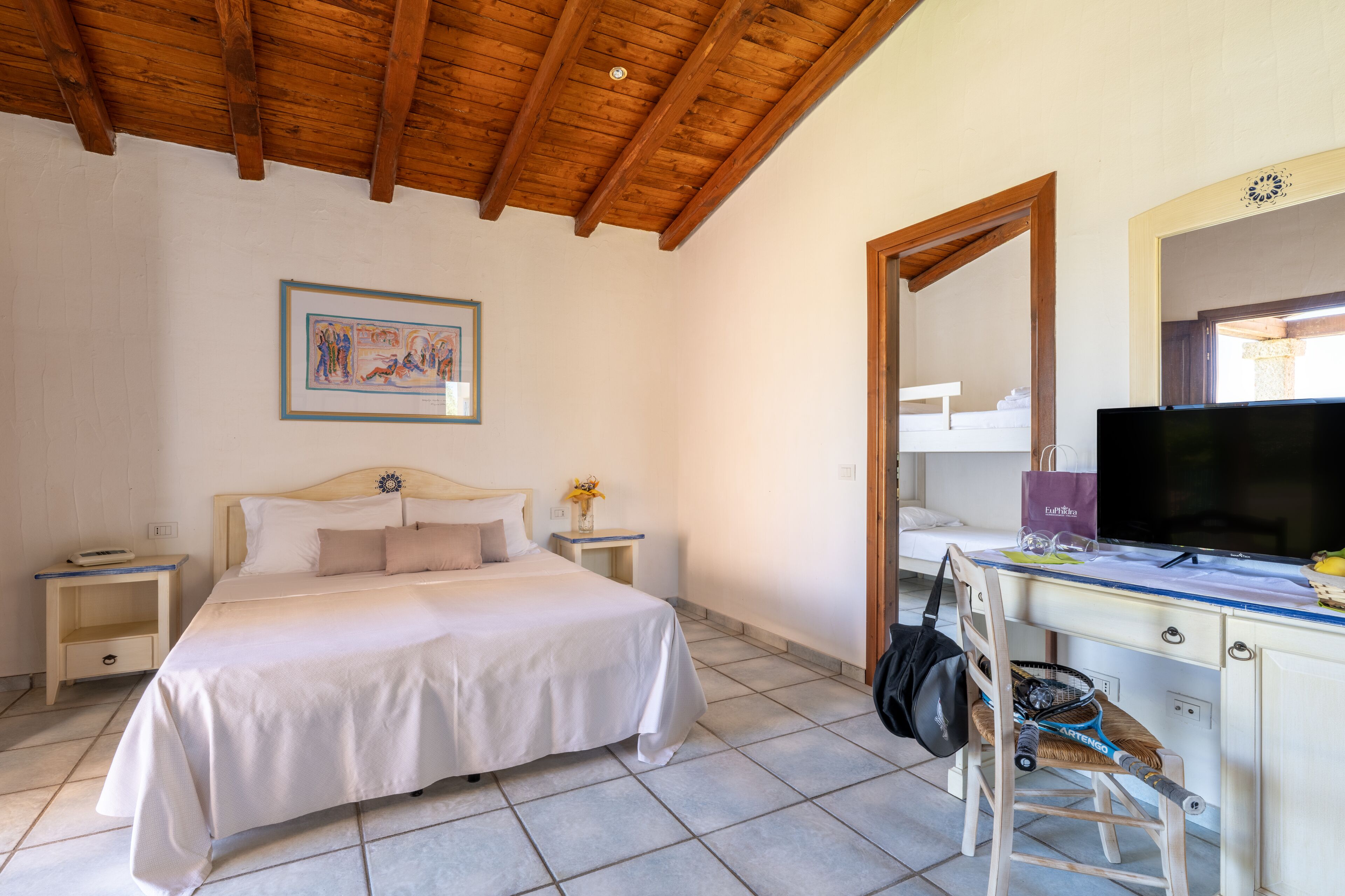 Un dormitorio con una cama, mesitas de noche, un escritorio con silla, televisor y espejo.
