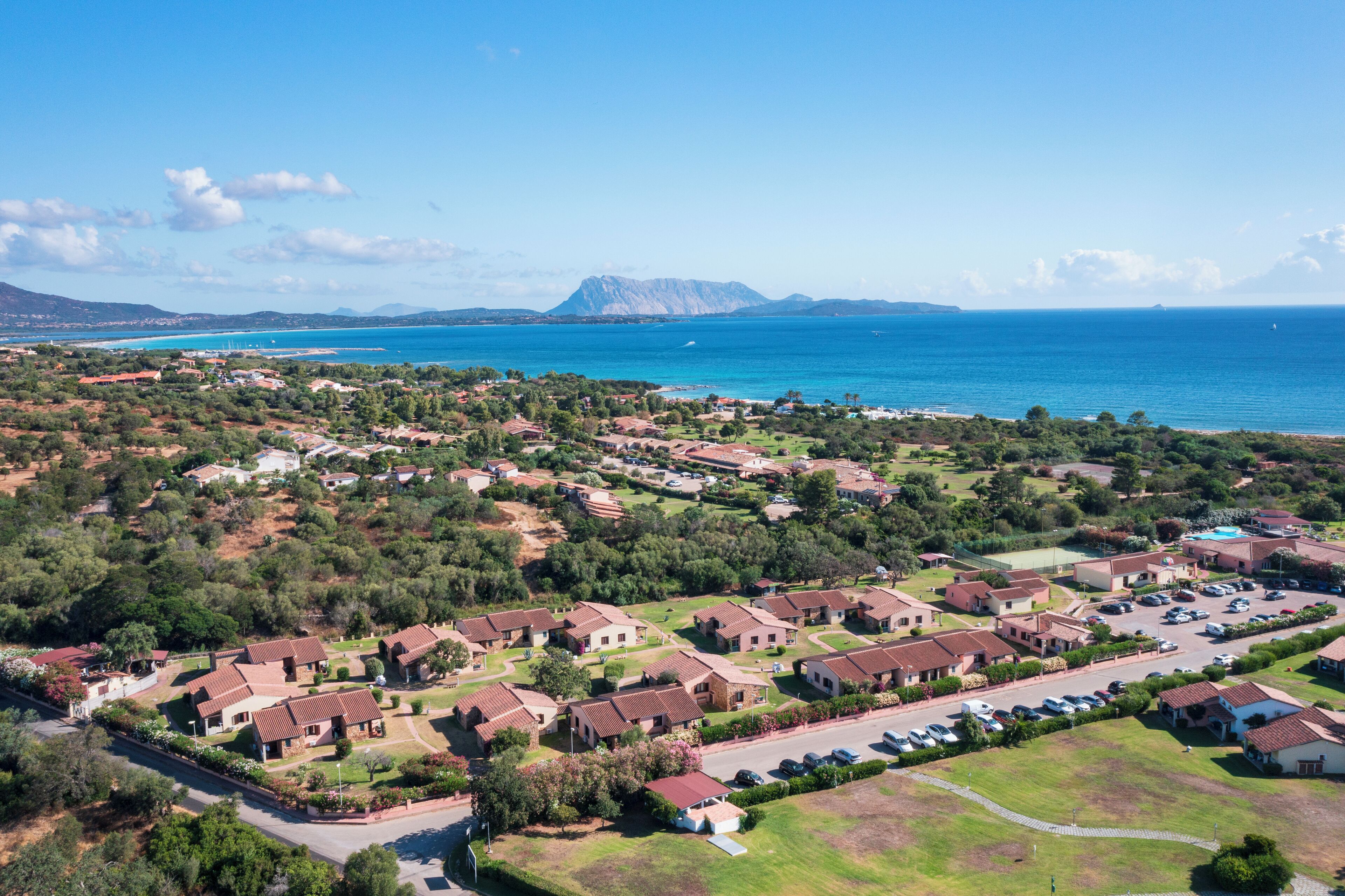 An aerial view of a coastal residential area with houses, a road, and a large body of water in the background.