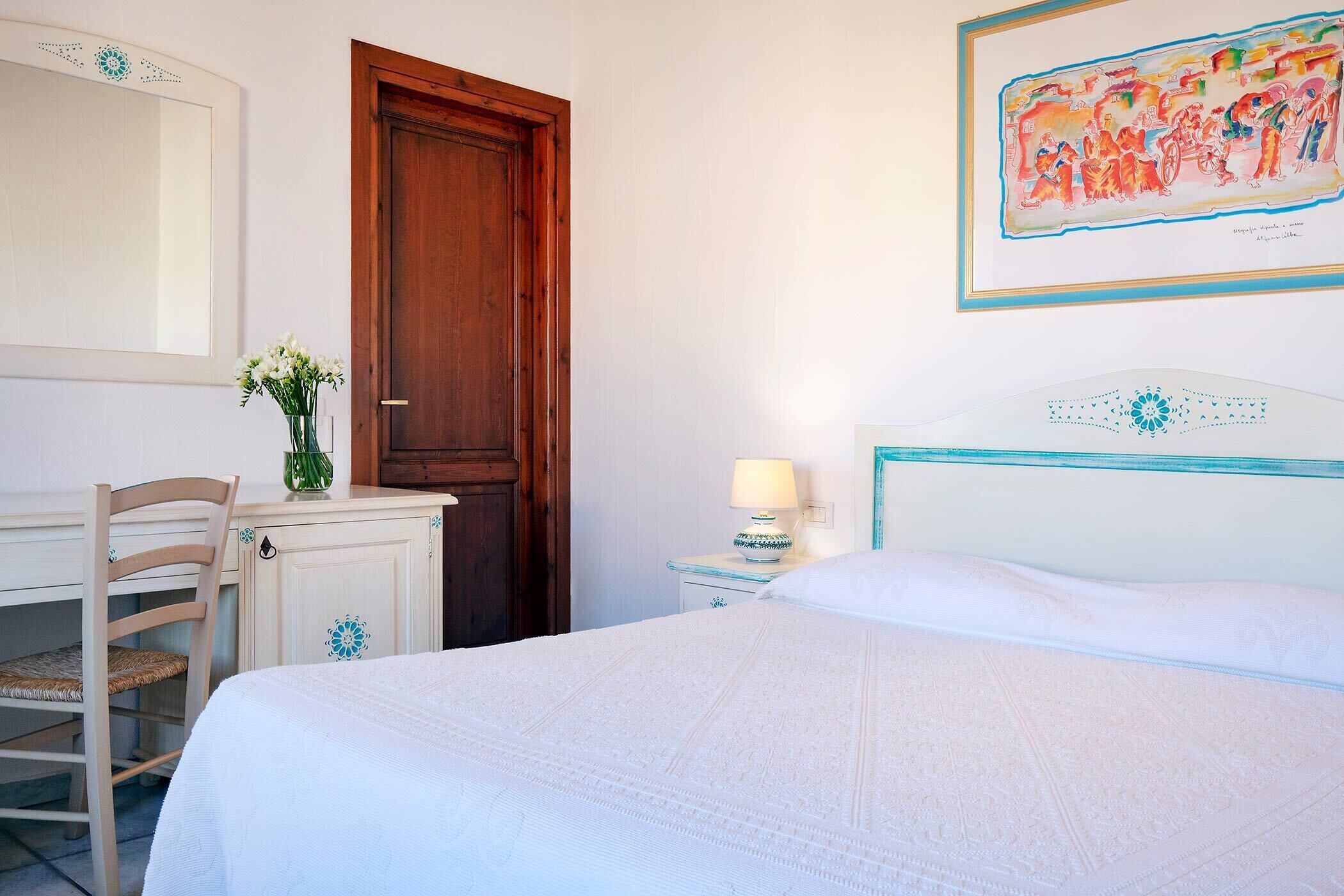 A bedroom with a bed, a desk, a chair, a wooden door, a nightstand with a lamp, and a wall with a framed artwork.