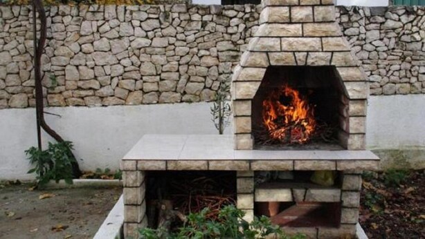 A stone-built outdoor fireplace with a fire burning inside, set against a stone wall and a white building in the background. - Apartments Sikirica 1 (Trogir)