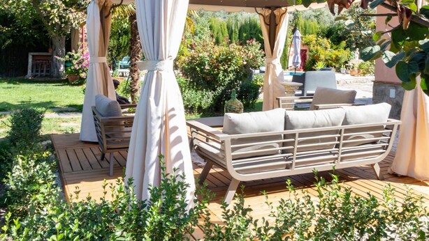 Et overdækket udendørs opholdsområde med pergola, træterrasse og frodig beplantning.