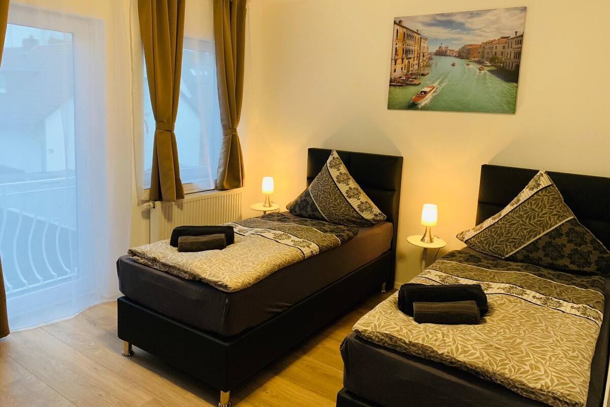 Une chambre d’hôtel avec deux lits, une fenêtre avec des rideaux, un radiateur, une lampe fixée au mur et un tableau représentant un paysage de canal.