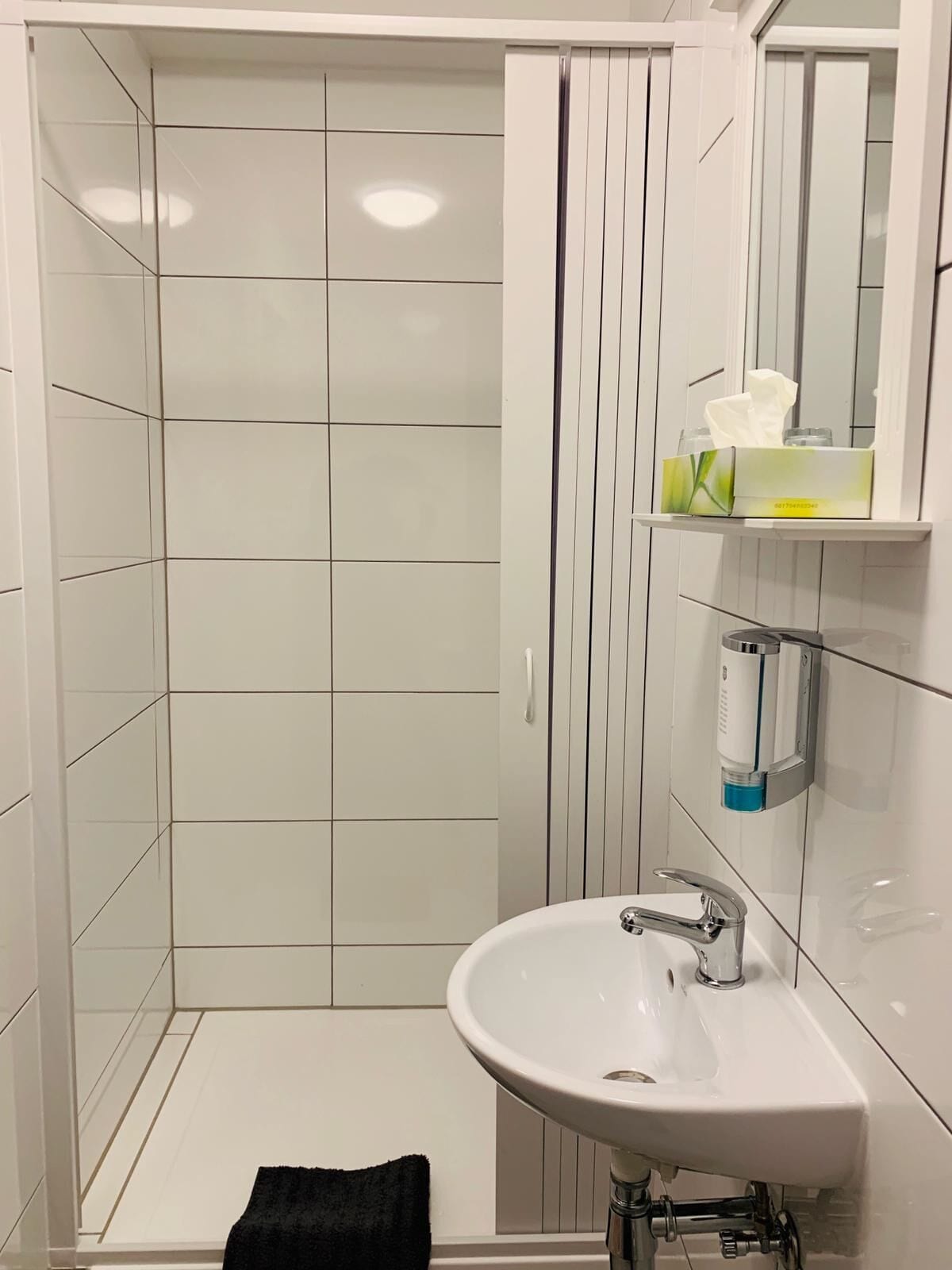 Un baño con ducha de azulejos blancos, un lavabo con espejo y un estante con artículos de aseo.