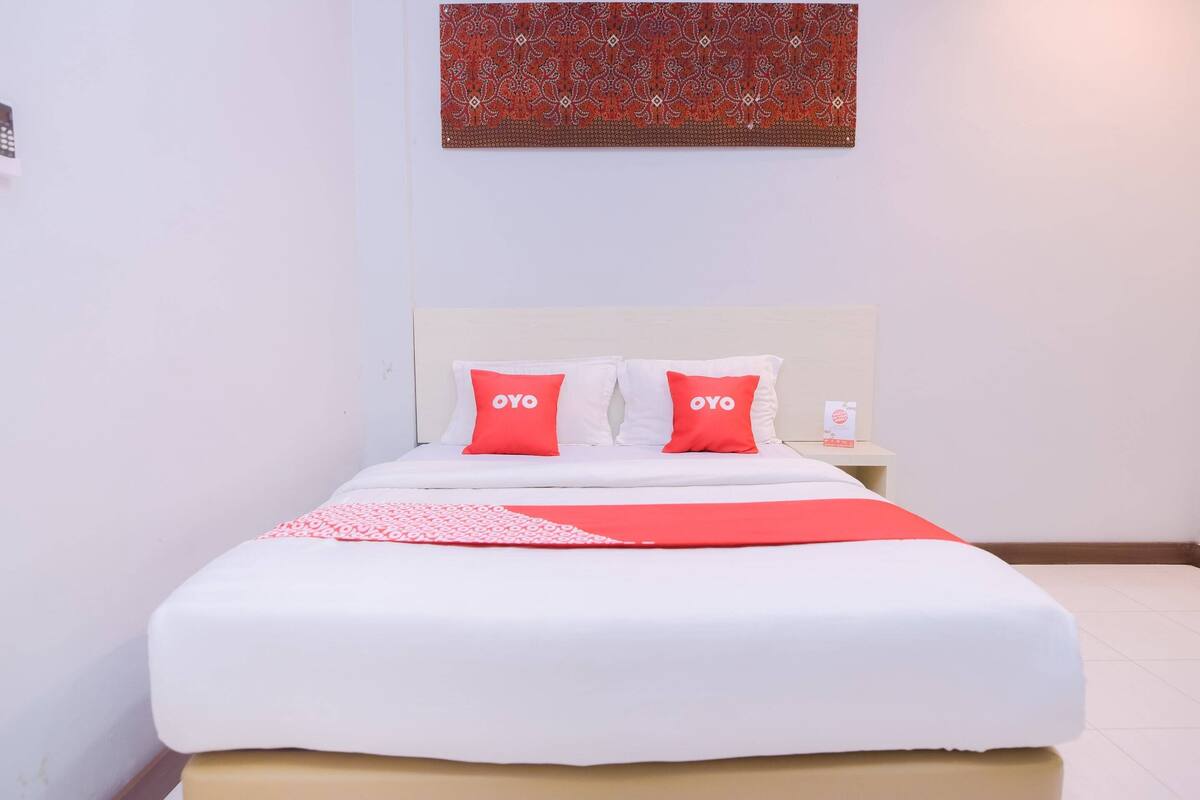 A neatly made bed with white and red bedding, a patterned wall hanging above, and a small bedside table with a lamp.