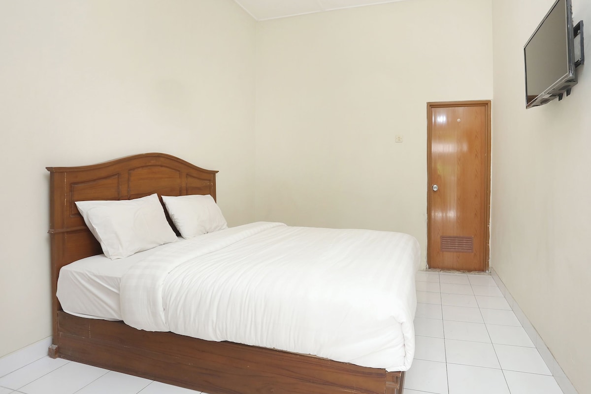 Une chambre avec un lit en bois, du linge de lit blanc, un téléviseur à écran plat fixé au mur et une porte dotée d’un judas.