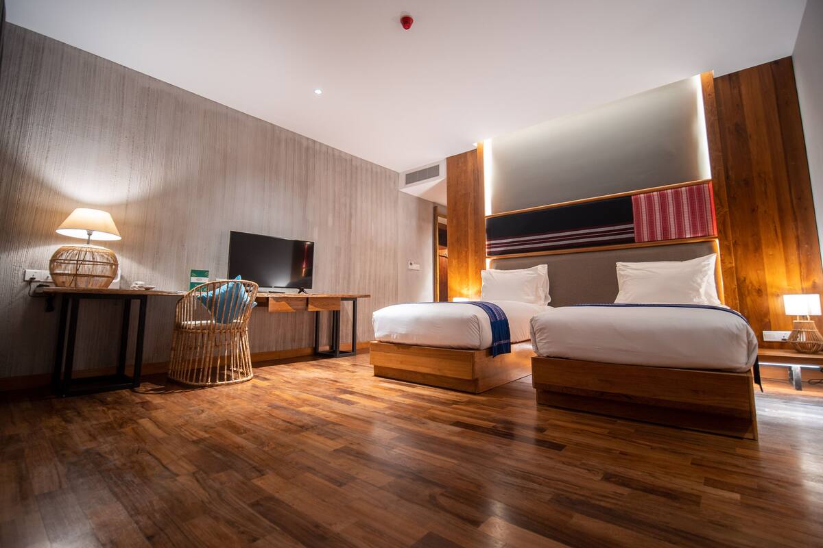 A hotel room with a wooden floor, a desk with a lamp, a television, a bed with two pillows, and a headboard with a striped design.
