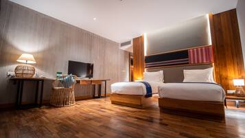 A hotel room with a wooden floor, a desk with a lamp, a television, a bed with two pillows, and a headboard with a striped design.