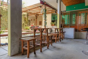 Un espacio al aire libre techado con mesas y sillas de madera, una planta colgante y una pared verde con arte de bambú.