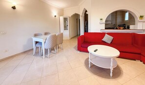 A living room with a red sofa, a white coffee table, a dining table with chairs, and a kitchen visible through an archway.