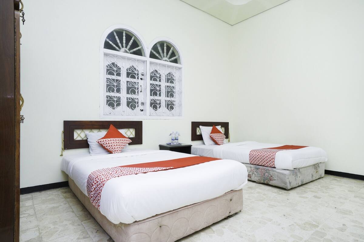 A hotel room with two beds, a window with decorative grills, a wooden headboard, and a tiled floor.