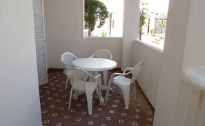 Uma pequena varanda com uma mesa branca e quatro cadeiras brancas, piso estampado e vista para um jardim e edifícios.
