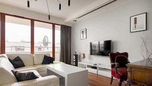 A modern living room with a white sofa, a coffee table, a red armchair, a TV, and a large window with a view of buildings. - Hlonda Deluxe Apartment with Terrace (Warsaw)