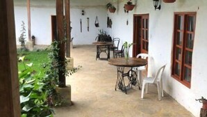 Une terrasse couverte avec un sol en pierre, une table entourée de chaises, des plantes suspendues et une vue sur le jardin.