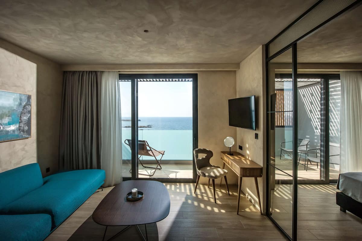 Una habitación de hotel moderna con sofá, escritorio, silla, televisor y balcón con vista al mar.
