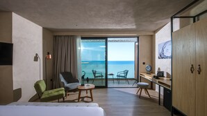 Un cuarto de hotel con una cama, un sillón verde, una mesa redonda, un escritorio con una silla y vista al mar por una puerta corrediza.