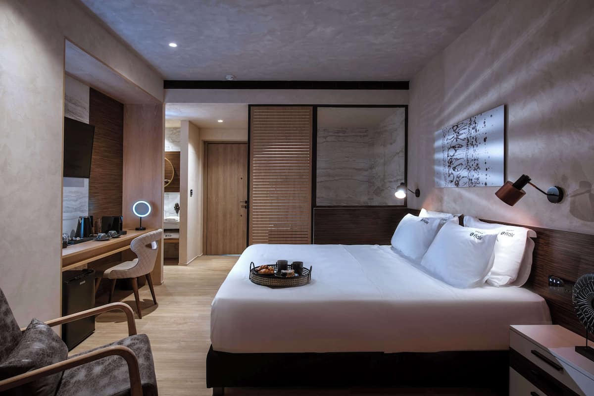 Una habitación de hotel moderna con una cama grande, un escritorio con silla, una mesita de noche y una lámpara de pared.