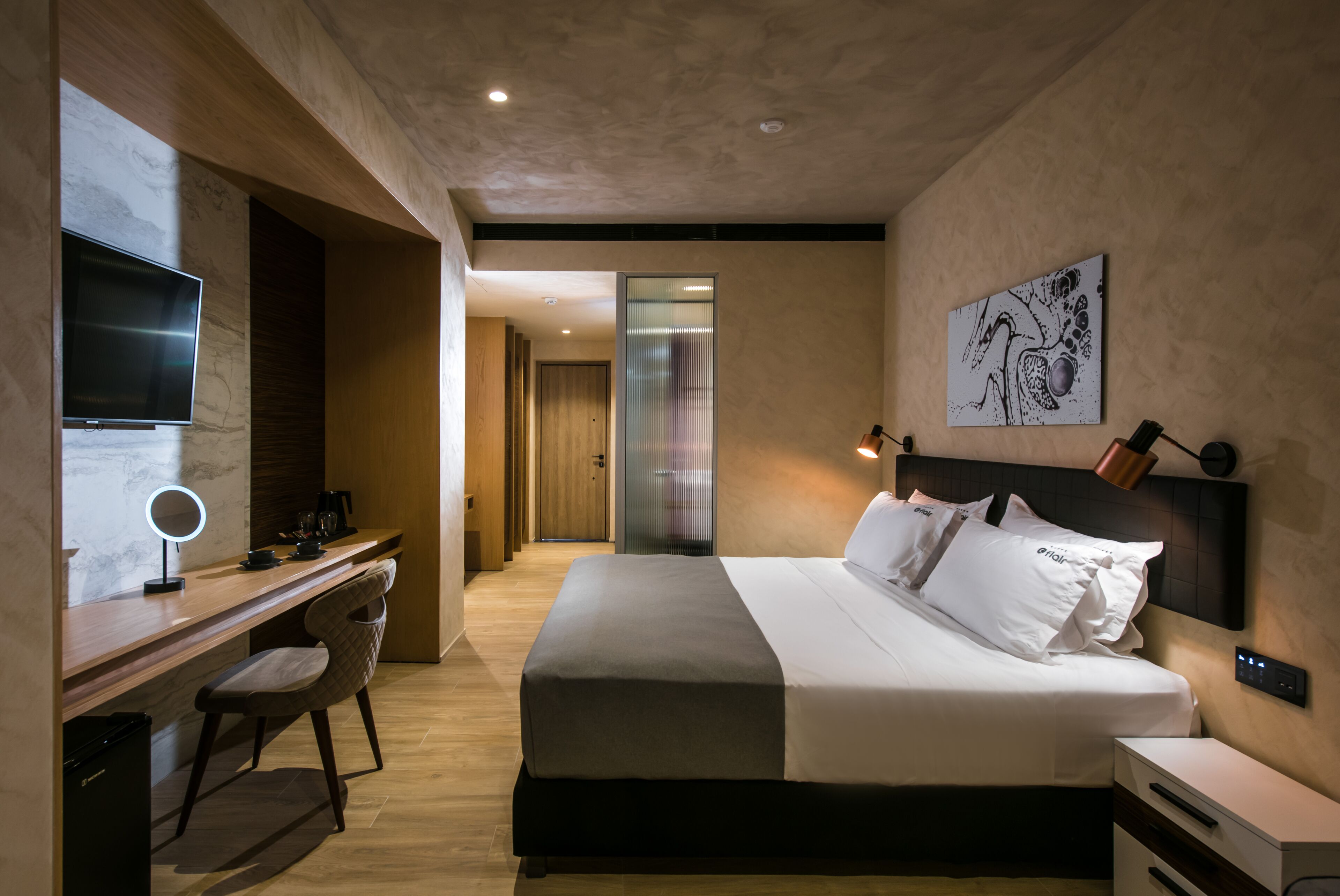 A modern hotel room with a large bed, a desk with a computer and a lamp, and a TV on the wall.