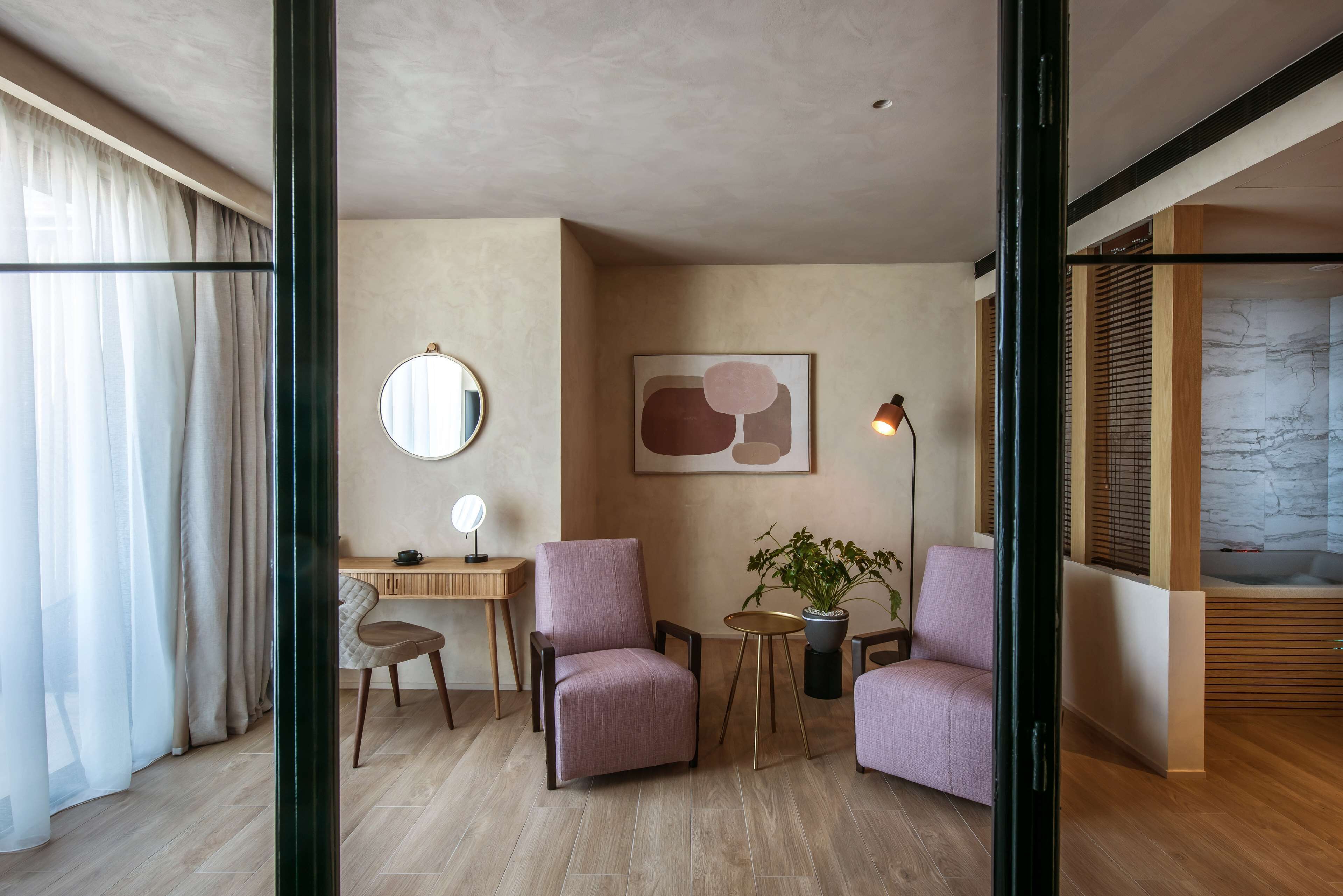 Habitación de hotel moderna con suelo de madera, un escritorio con una silla, dos sillones, un espejo redondo, una planta en maceta y un ventanal con cortinas.