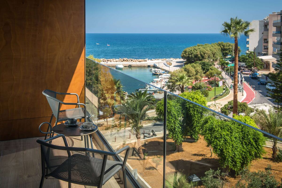 Un balcón con mesa y sillas que da a un puerto deportivo con barcos, una playa y un paisaje urbano.