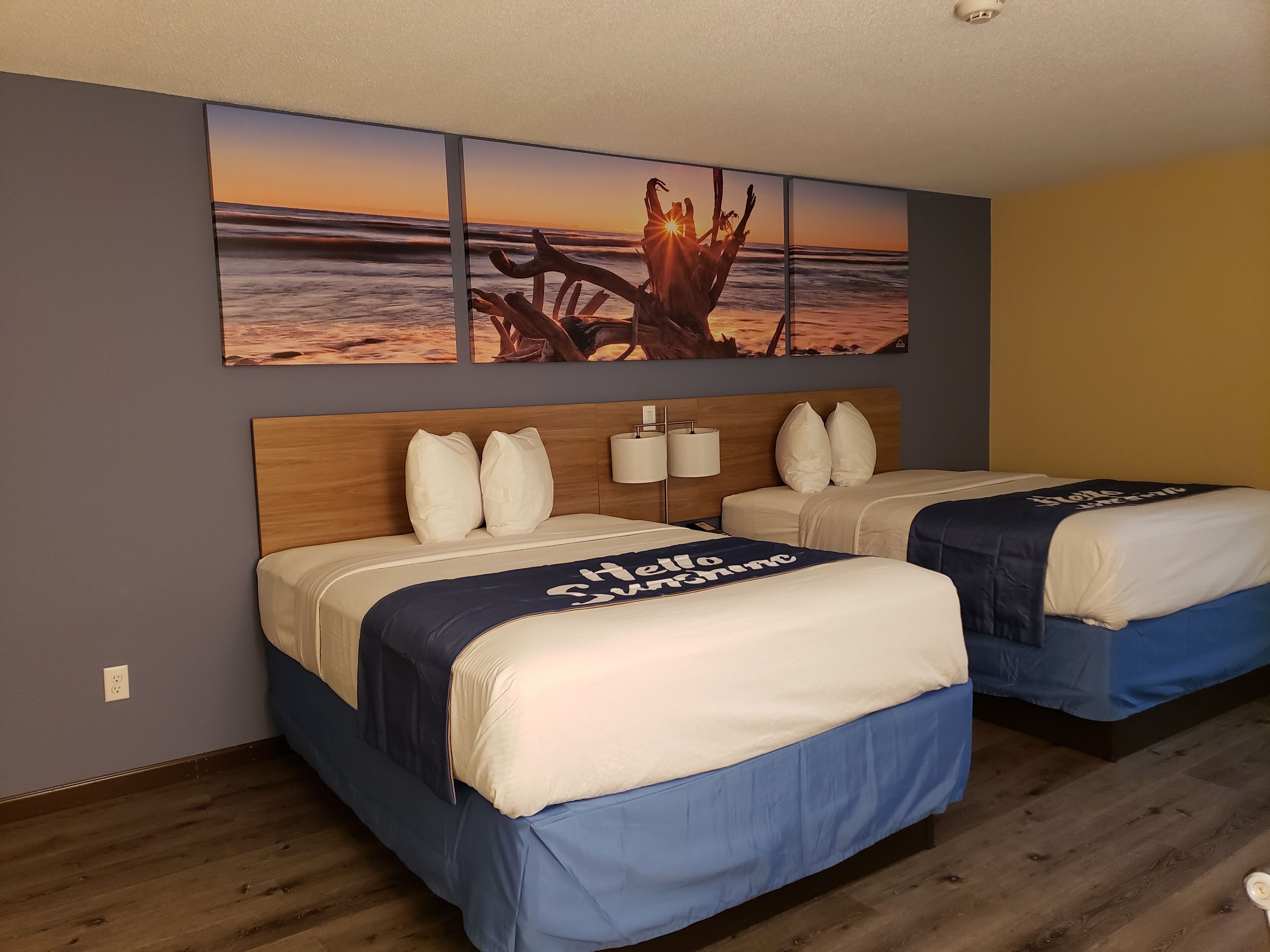 Habitación de hotel con dos camas, cabecera de madera, lámpara de pared y un mural con vista a la playa al atardecer.