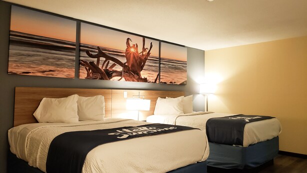 A hotel room with two beds, a large wall mural of a sunset over the ocean, and a nightstand with a lamp. - Days Inn & Suites by Wyndham Merrillville (Merrillville)