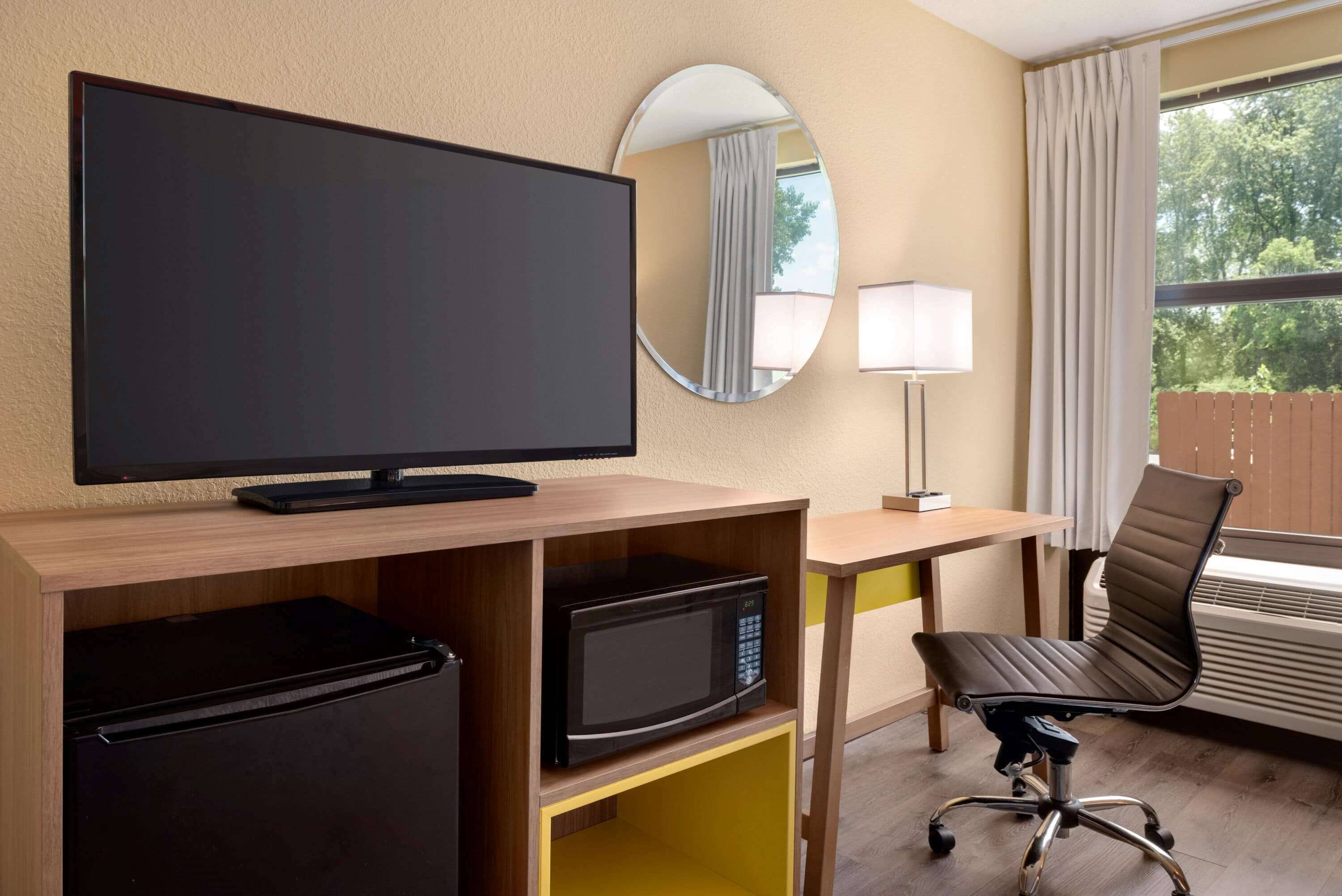 A hotel room with a flat-screen TV, a microwave, a mini-fridge, a desk with a lamp, a chair, and a large window with curtains.