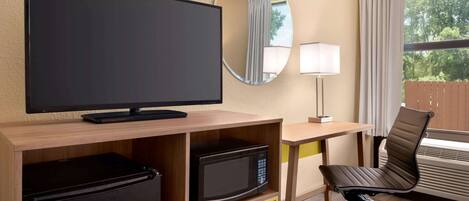 A hotel room with a flat-screen TV, a microwave, a mini-fridge, a desk with a lamp, a chair, and a large window with curtains.
