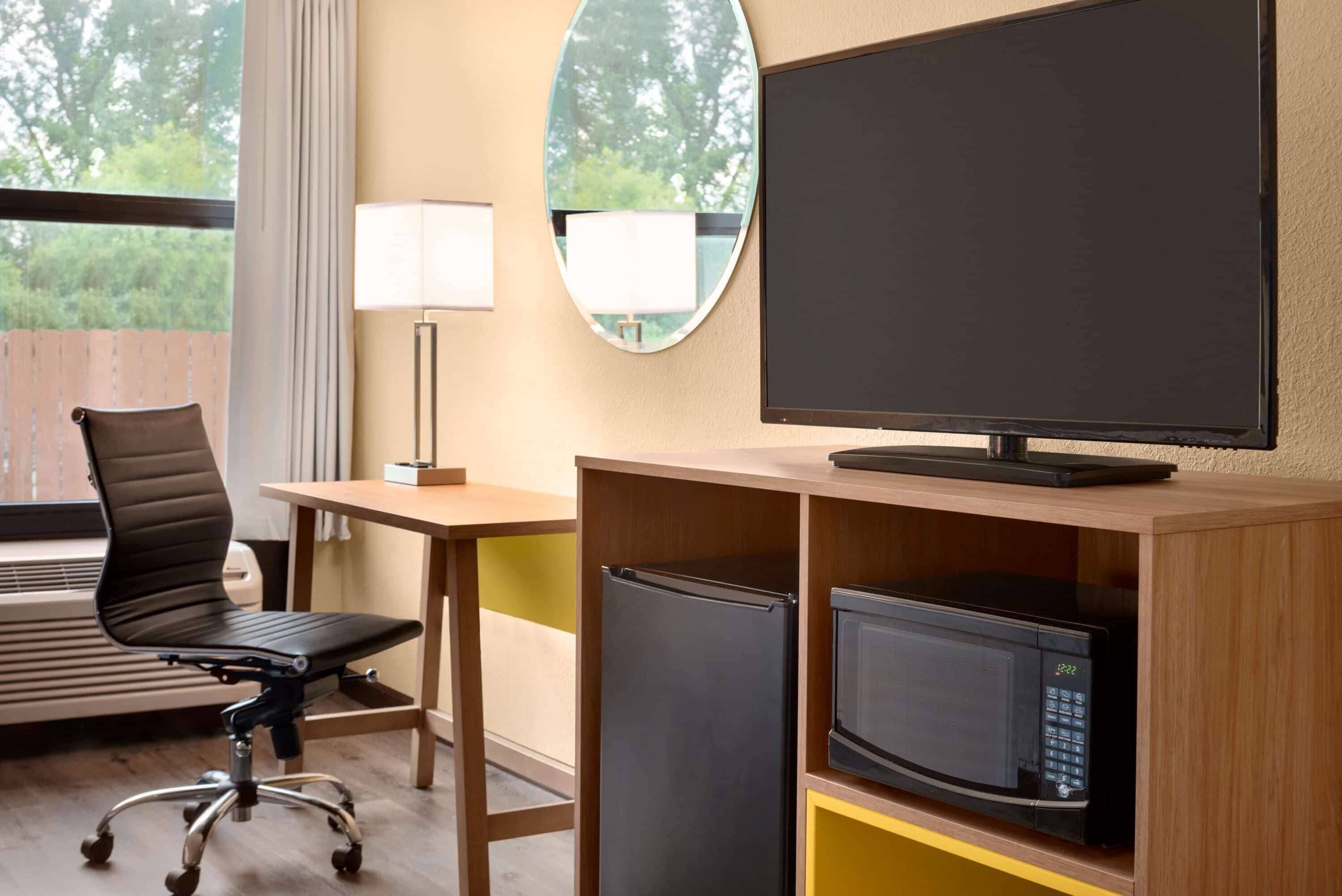 A hotel room with a wooden desk, a black office chair, a flat-screen TV, a microwave, and a small refrigerator.