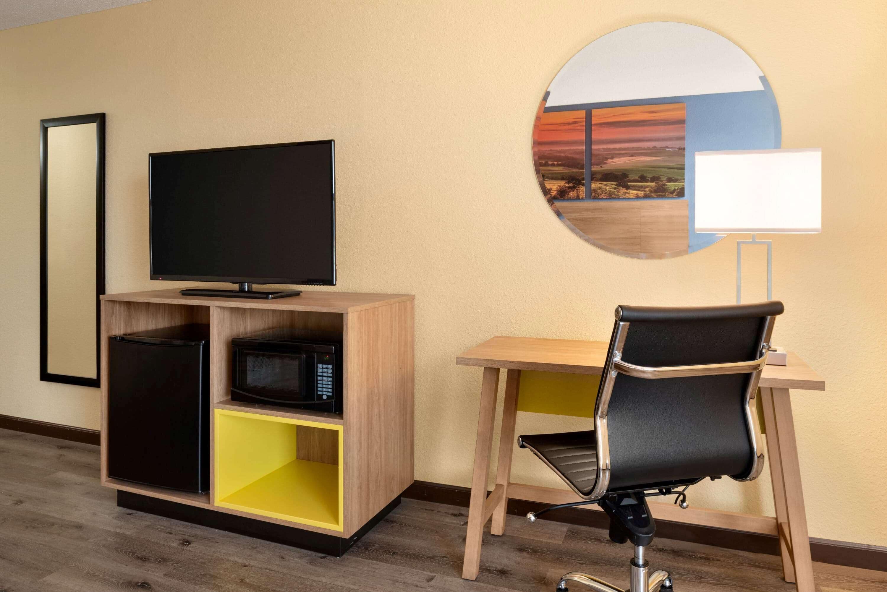 A hotel room with a television, a microwave, a desk with a chair, and a wall-mounted lamp.
