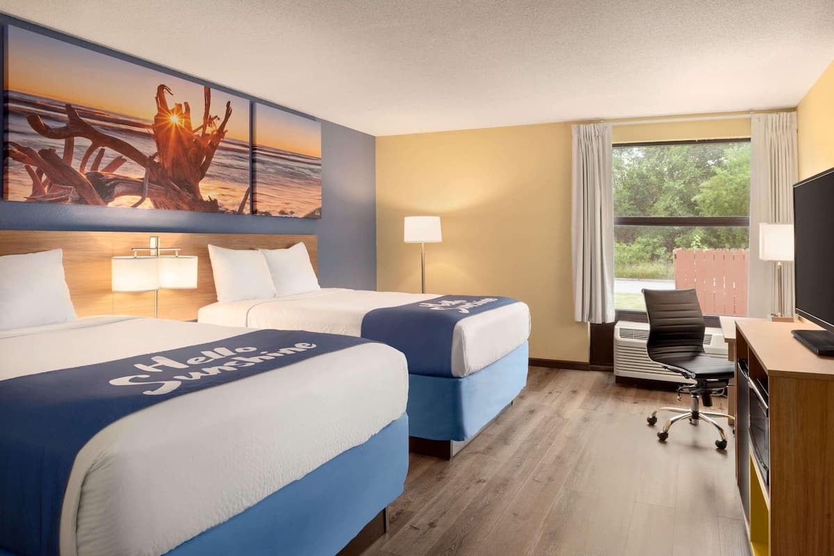 A hotel room with two beds, a desk, a chair, a TV, and a painting of a sunset over the ocean.