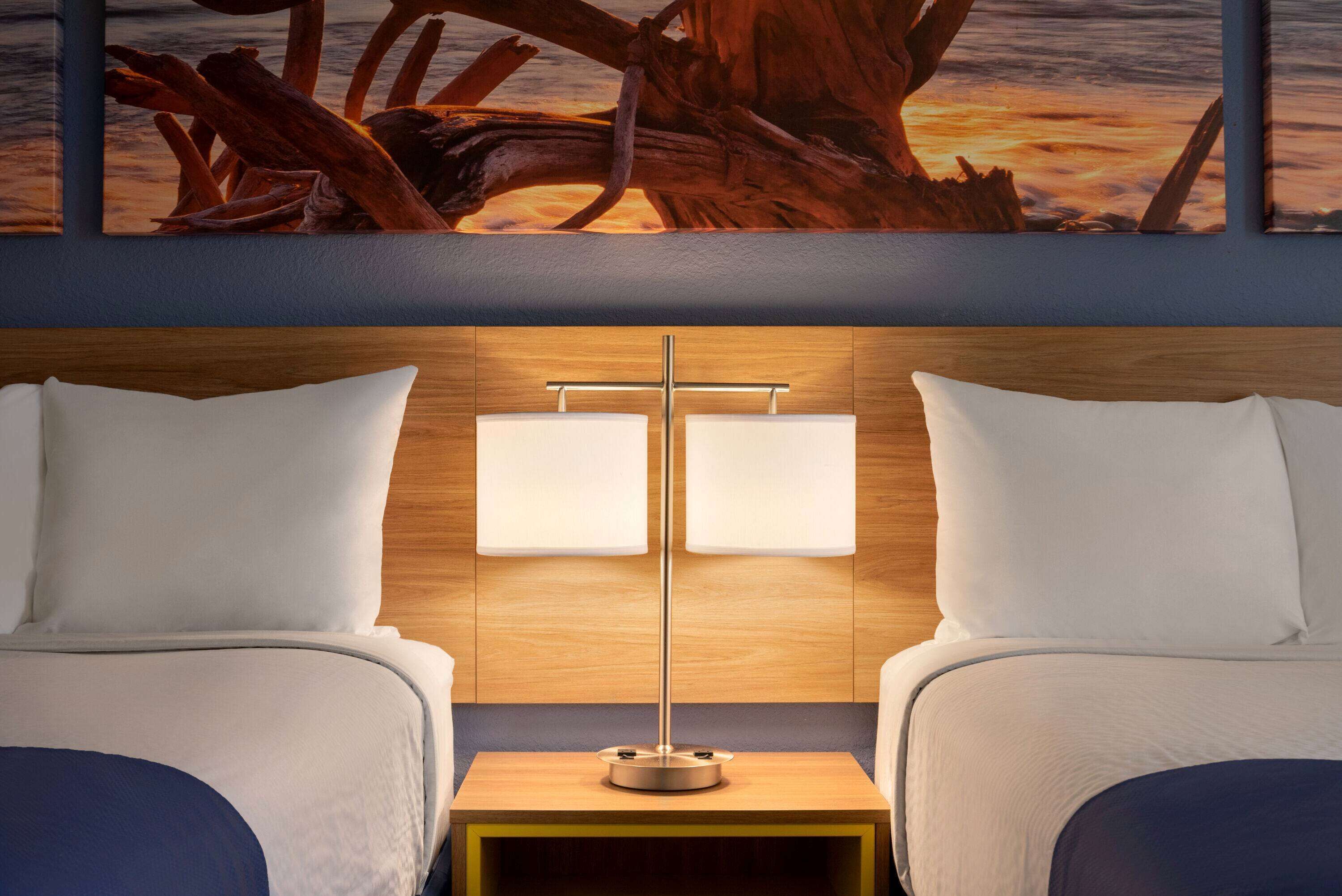 A hotel room with two beds, a nightstand with a lamp, and a large painting above the beds.