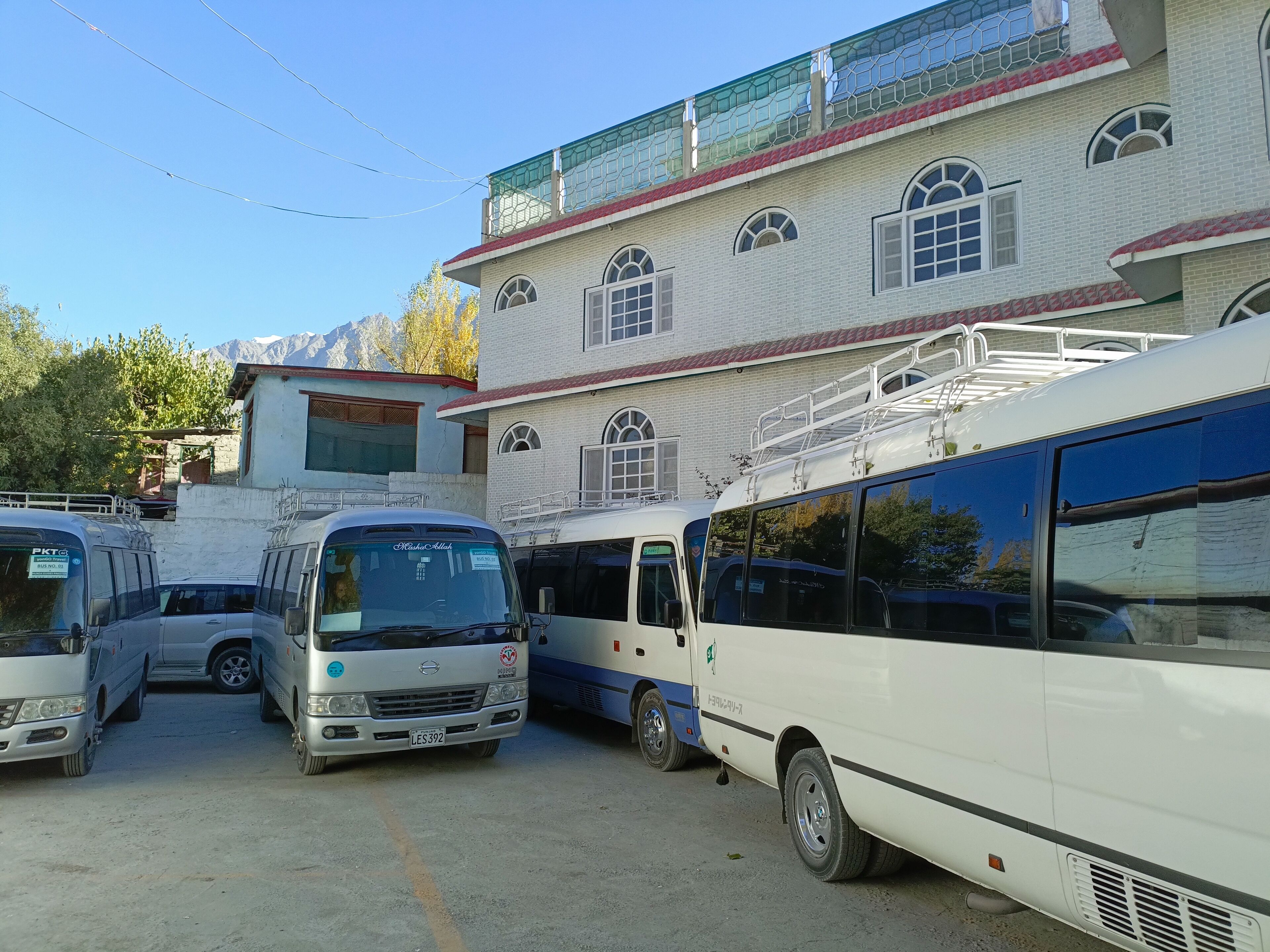 Un estacionamiento con varios autobuses, un edificio de múltiples ventanales y techo rojo, y montañas al fondo.