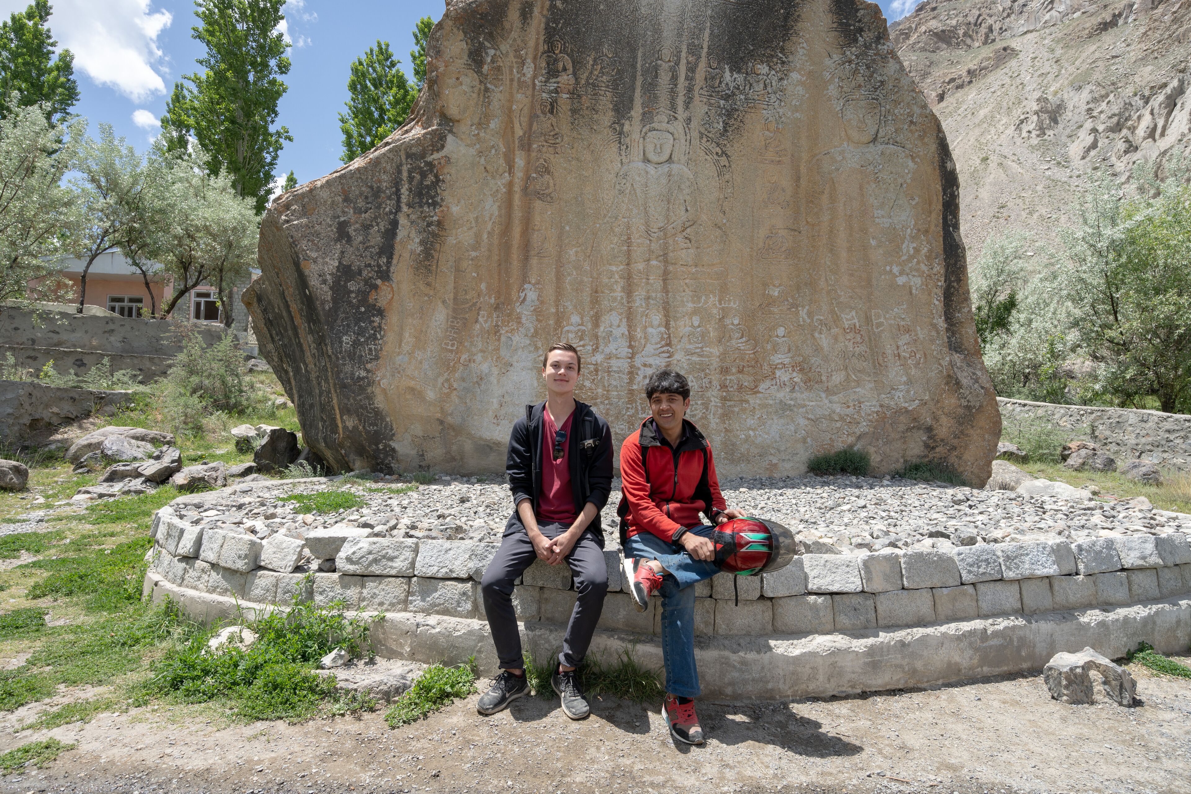 Two people sitting on a stone bench in front of a large rock with carvings, surrounded by a stone wall and greenery.