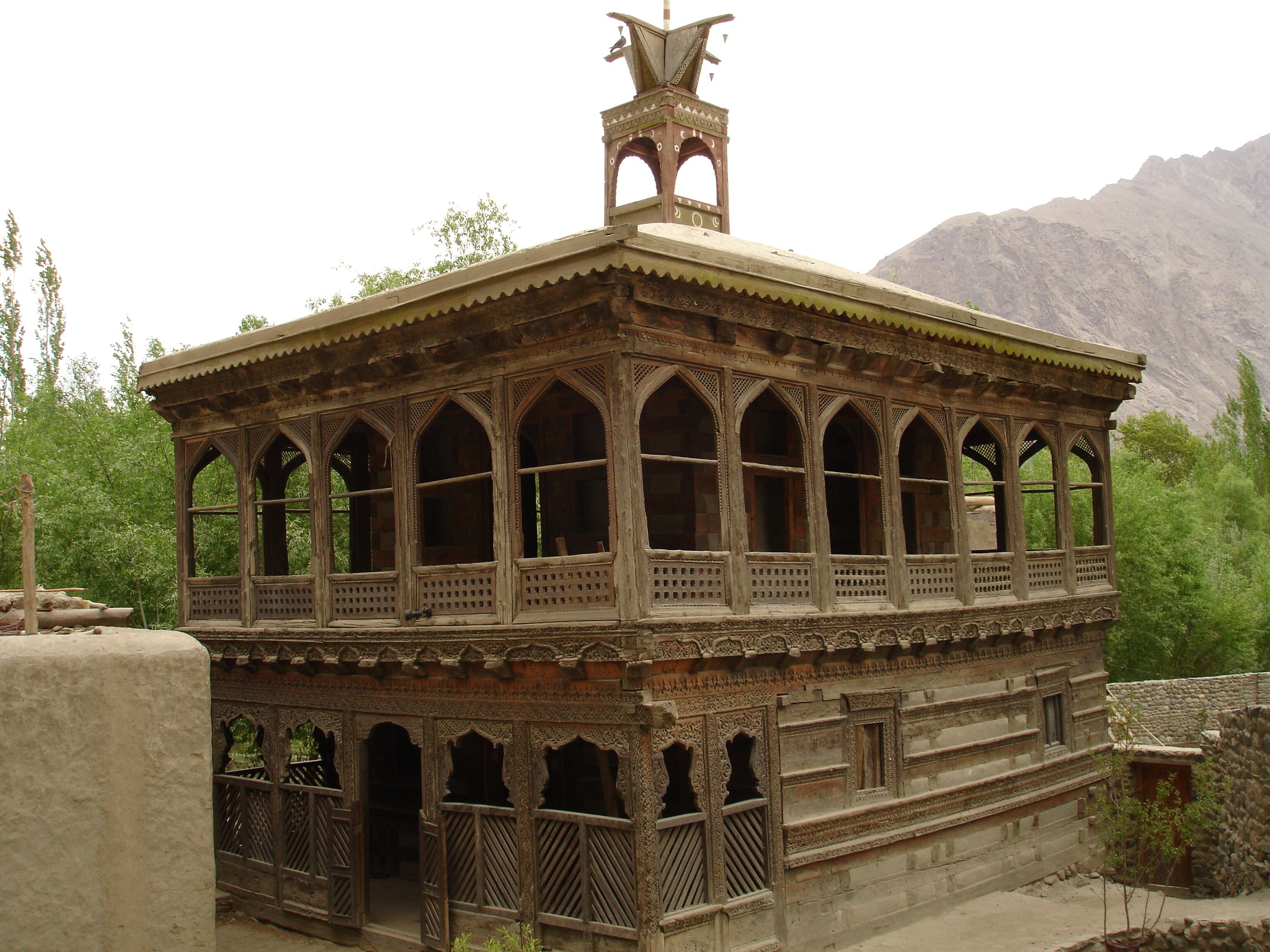 Una estructura de madera tradicional con balcón y torre, rodeada de vegetación y montañas.