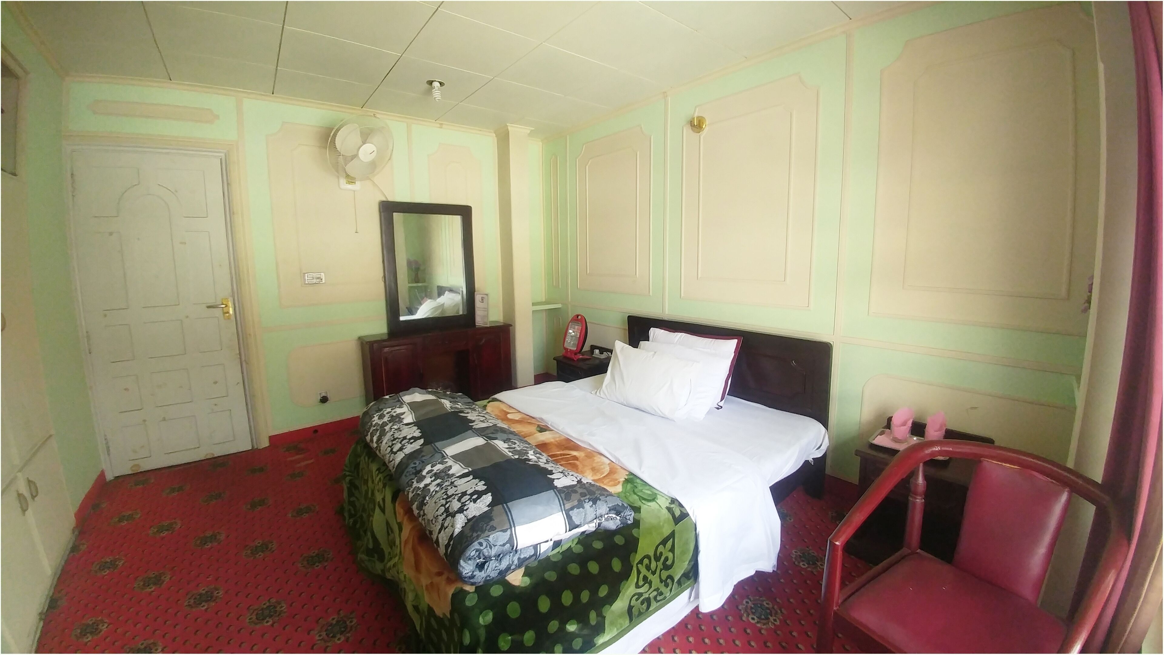 A hotel room with a bed, a nightstand, a mirror, a red chair, and a door.