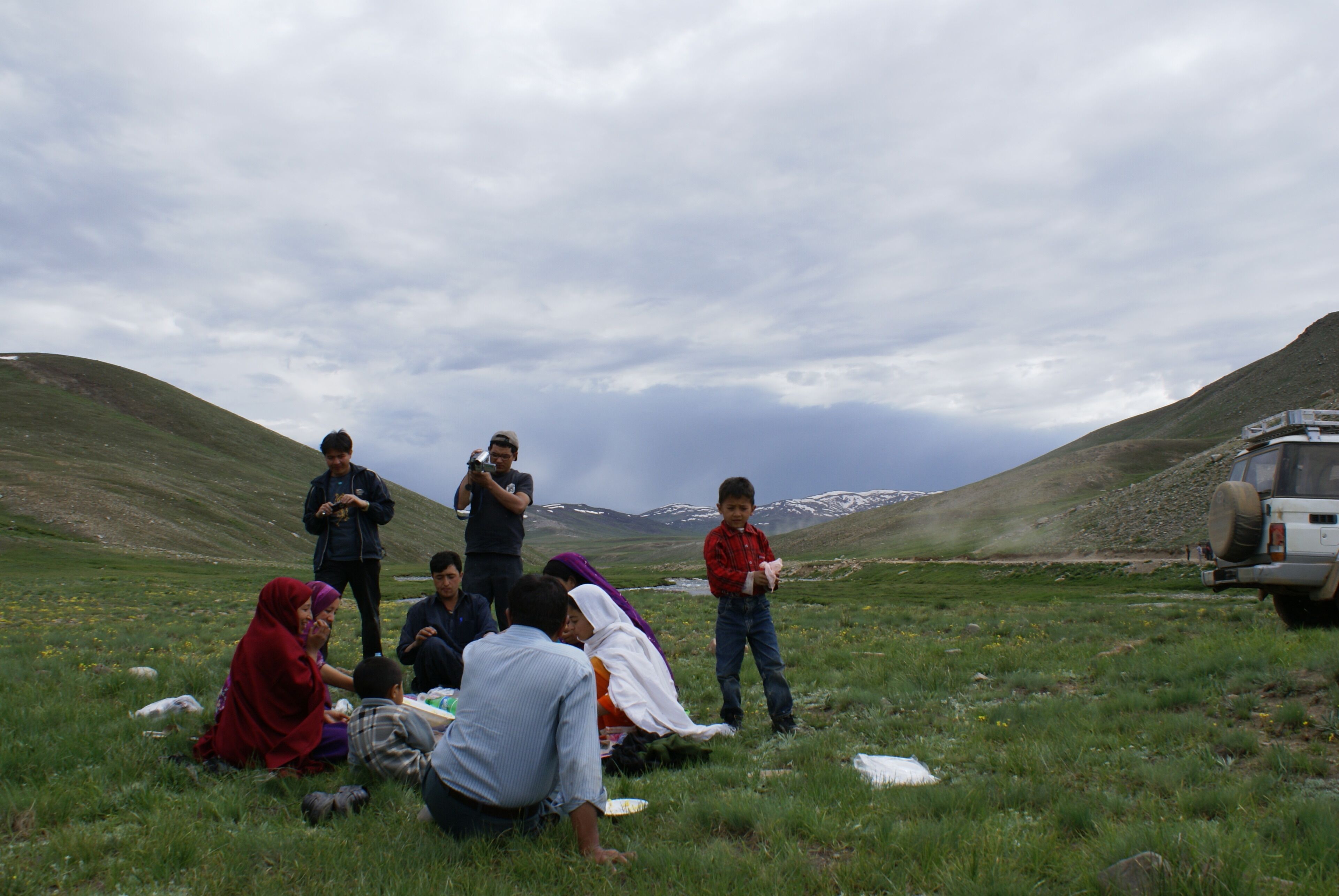Un grupo de personas sentadas sobre la hierba en una zona montañosa, con un vehículo al fondo.