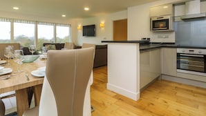 A modern kitchen with a built-in oven, a dining area with a table set for four, and a seating area with a sofa. - Juniper (Truro)