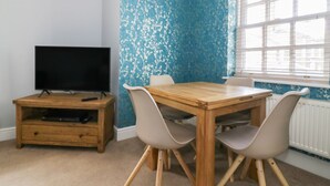 A room with a wooden table and four chairs, a television on a wooden stand, and a window with blinds.